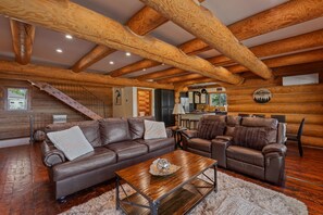 A spacious living area with a wooden ceiling and exposed beams, a leather sofa, a coffee table, and a kitchen in the background. - Bottle Bay Beauty | Lake Views Near Sandpoint (Sagle)