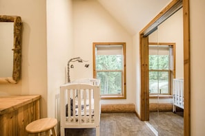 A room with a crib, a wooden chair, a large mirror, and two windows with a view of trees. - Bottle Bay Beauty | Lake Views Near Sandpoint (Sagle)