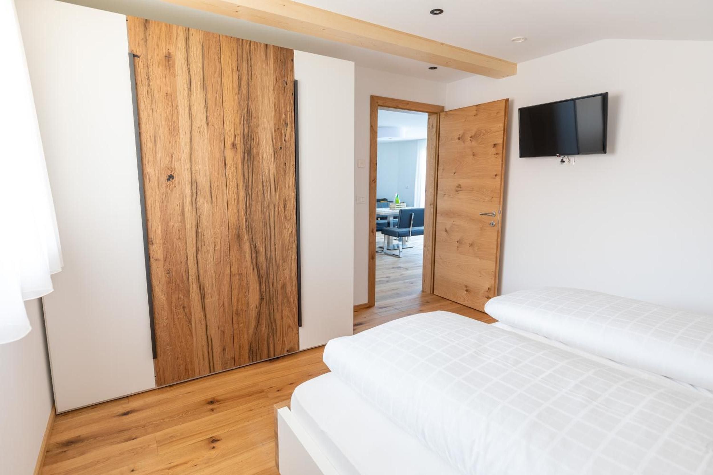 A bedroom with a wooden wardrobe, a bed with white bedding, and a television mounted on the wall.