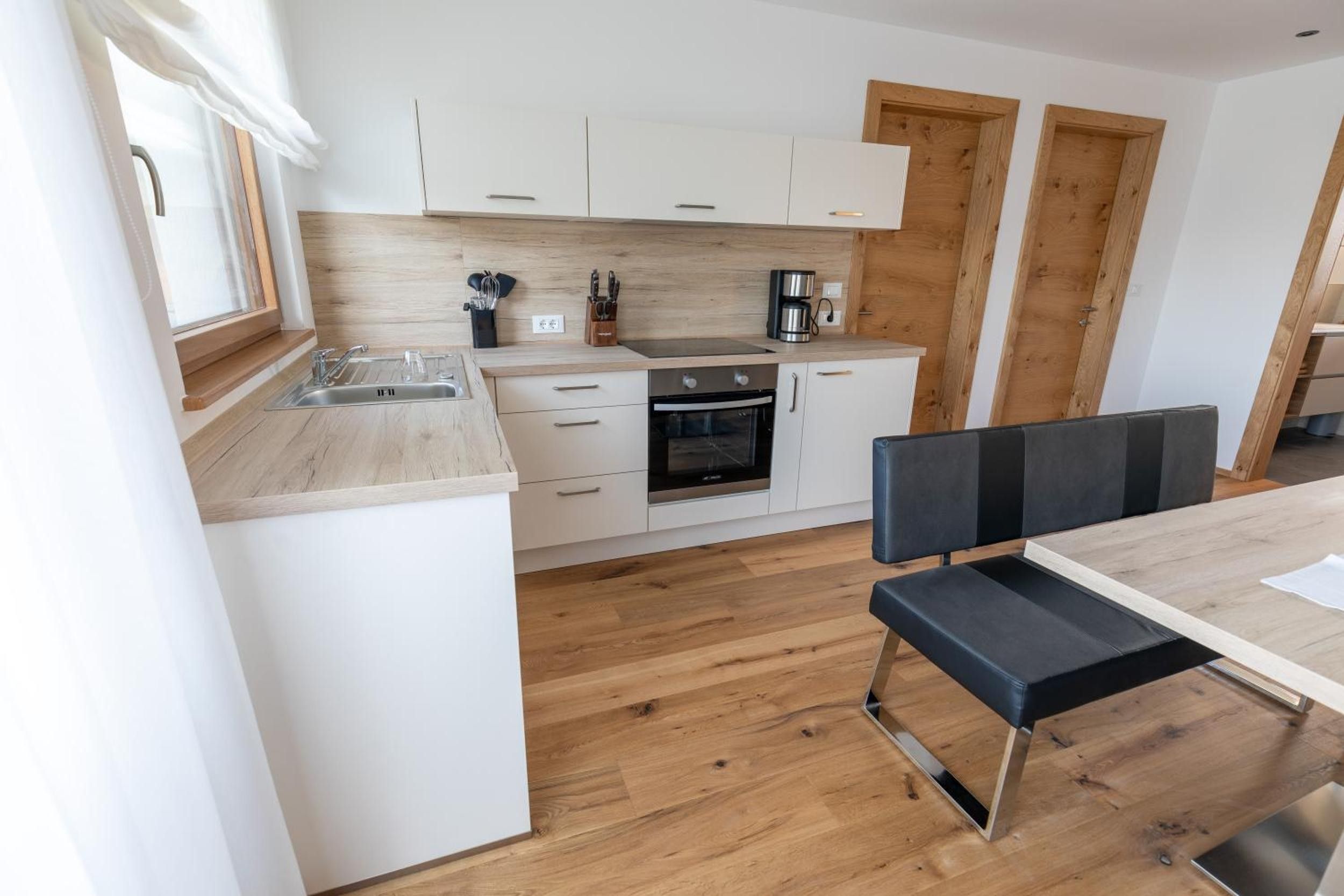 A modern kitchen with white cabinets, a stainless steel sink, a coffee maker, and a dining area with a bench and table.