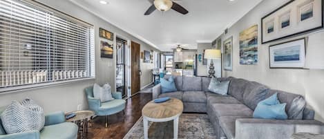 Un salon spacieux avec un grand canapé d’angle gris, une table basse ronde et deux fauteuils. On peut voir un ventilateur de plafond et des spots encastrés.
