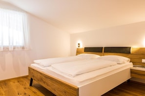 Ein Doppelbett mit weißen Leinen, ein hölzernes Kopfstück, ein Nachttisch mit Lampe und ein Fenster mit durchsichtigen Vorhängen.