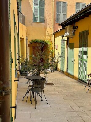 An outdoor seating area with tables and chairs, a small tree, and a building with green shutters.