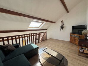 A loft-style room with a sloped ceiling, a sofa, a glass coffee table, a television on a wooden cabinet, and a skylight. - Duplex with terrace near beach (Merville-Franceville-Plage)