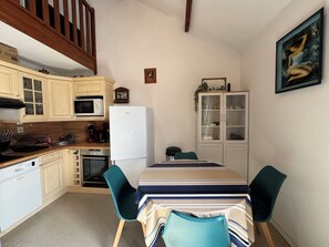 A compact kitchen with wooden cabinets, a white refrigerator, and a dining area with a striped tablecloth and teal chairs. - Duplex with terrace near beach (Merville-Franceville-Plage)