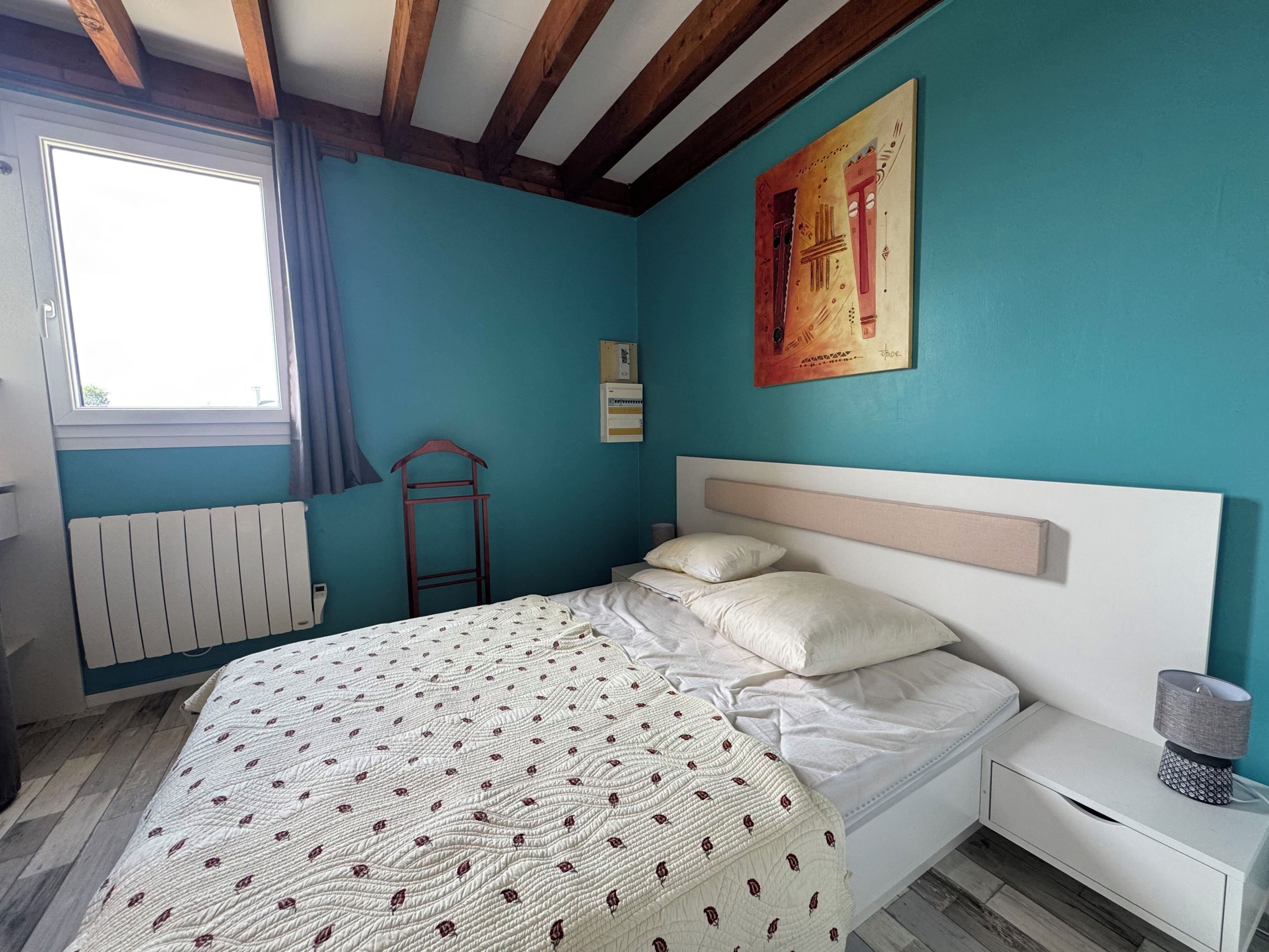 Un dormitorio con cama, mesita de noche, radiador, ventana y un cuadro en la pared.