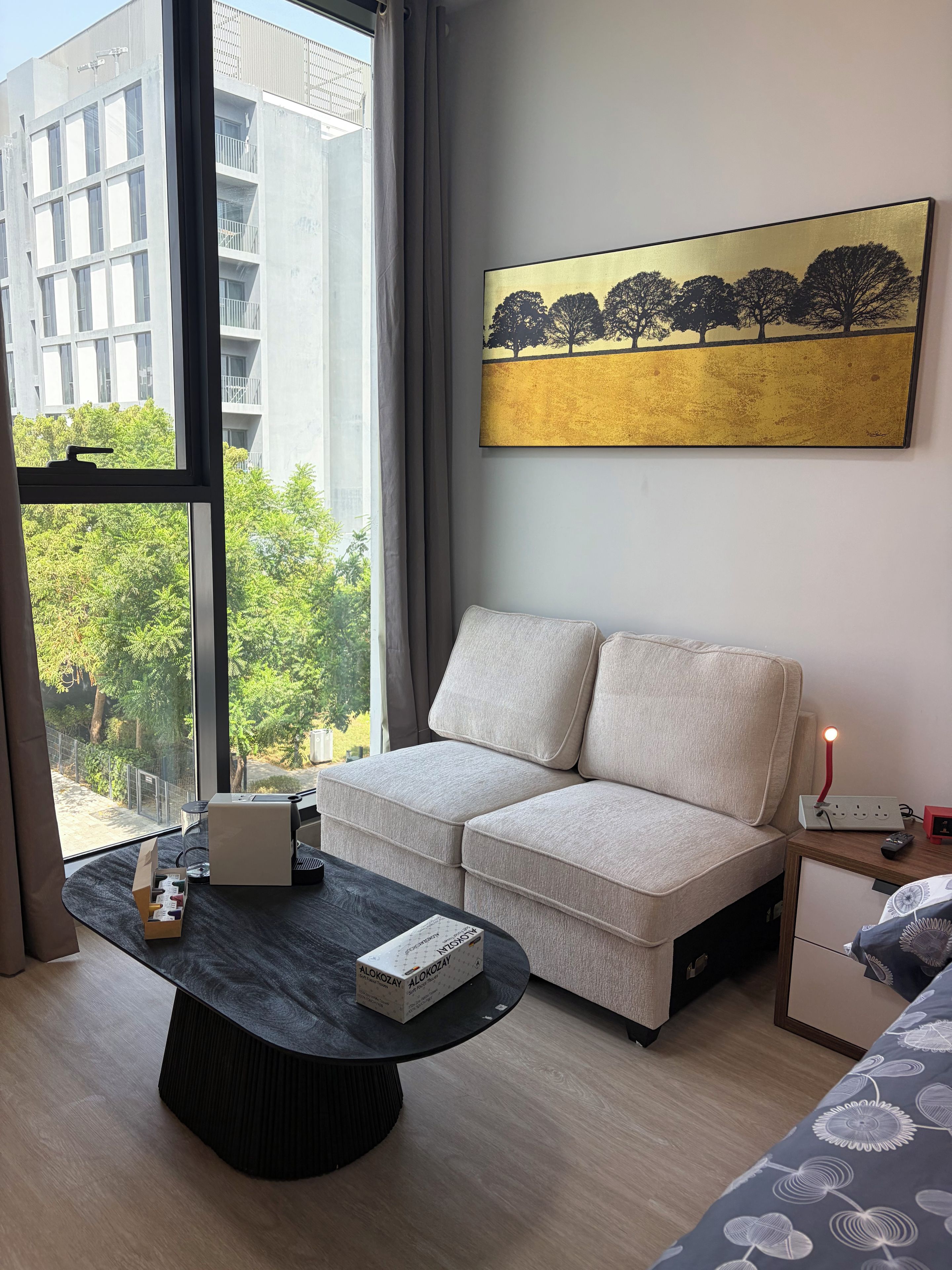 Ein modernes Hotelzimmer mit einer Couch, einem Couchtisch, einem Gemälde und Blick auf ein Gebäude und Bäume.