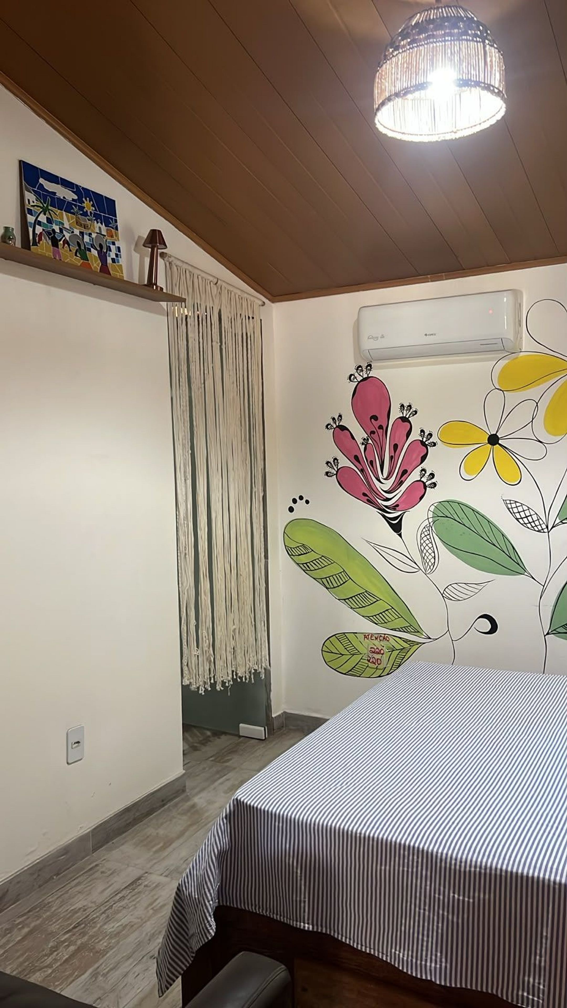Une chambre comprenant un lit, un luminaire suspendu, un appareil de climatisation et un mur orné d’une fresque florale aux couleurs vives.