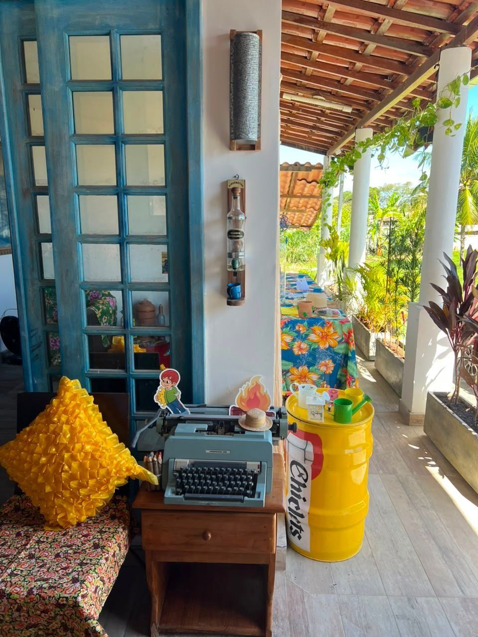 Una habitación con una puerta azul, una máquina de escribir, un barril amarillo y una mesa con mantel floral.