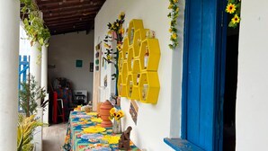 A covered outdoor area with a table covered in a floral tablecloth, a blue door, and a yellow hexagonal shelf with bottles. - Casa Velha Hospedagem Jacuípe (Camaçari)