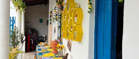 Área externa coberta com uma mesa adornada por uma toalha de mesa florida, uma porta azul e uma prateleira hexagonal amarela com garrafas.