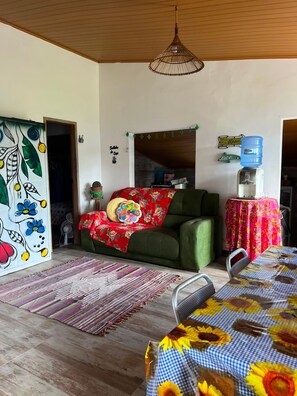 Una habitación con sofá, una mesa con mantel floral, dispensador de agua y una pared con letrero decorativo.