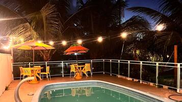 Una piscina exterior con sillas y sombrillas, rodeada de palmeras y una barandilla.
