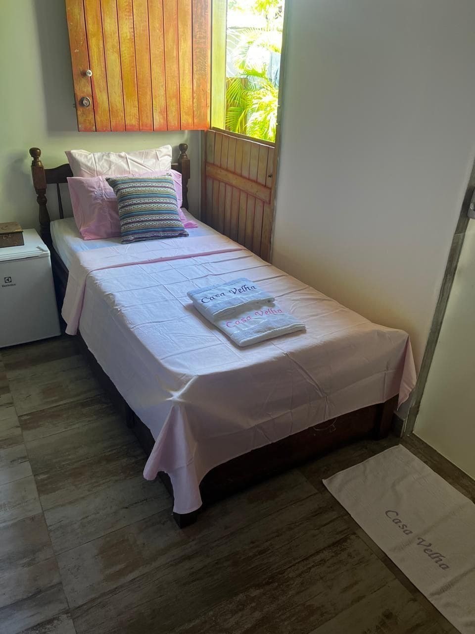 Un lit simple avec une couvre-lit blanche et une serviette ornée du texte « Casa Velha », une armoire en bois et un petit appareil électroménager blanc.