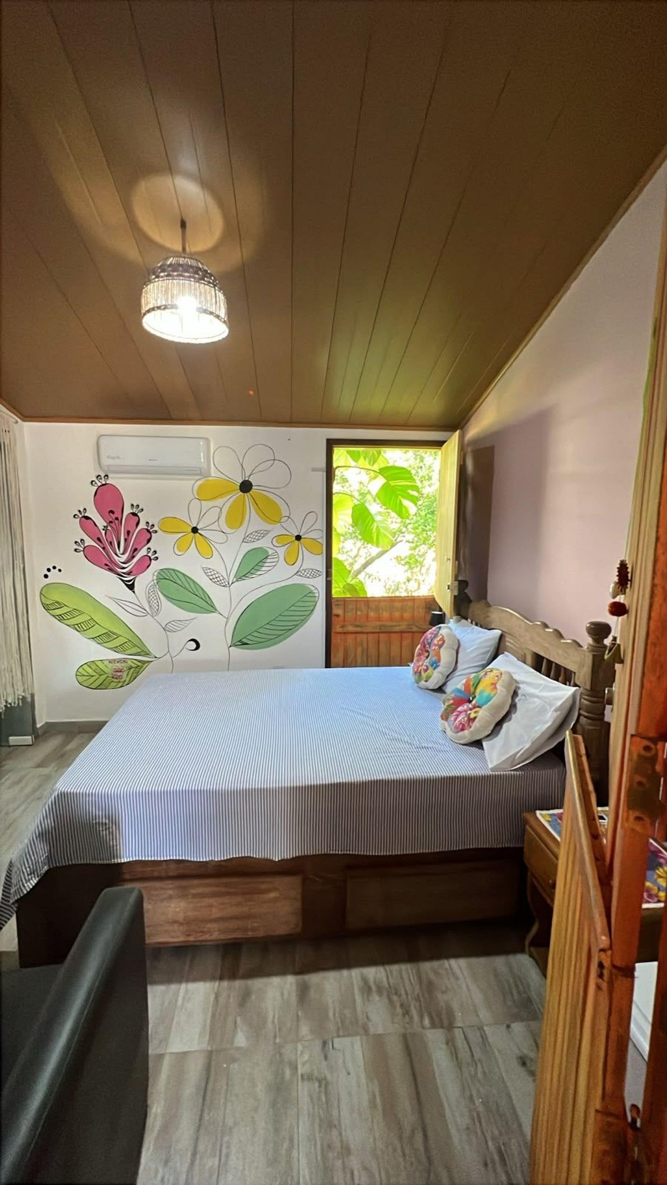 A bedroom with a bed, a wooden headboard, a nightstand, a wall with a floral mural, and a window with a view of greenery.