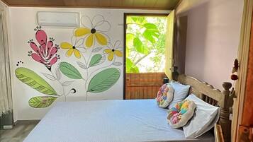 A bedroom with a bed, a wooden headboard, a nightstand, a wall with a floral mural, and a window with a view of greenery.