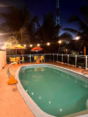 An outdoor swimming pool with chairs and umbrellas, surrounded by palm trees and a railing.