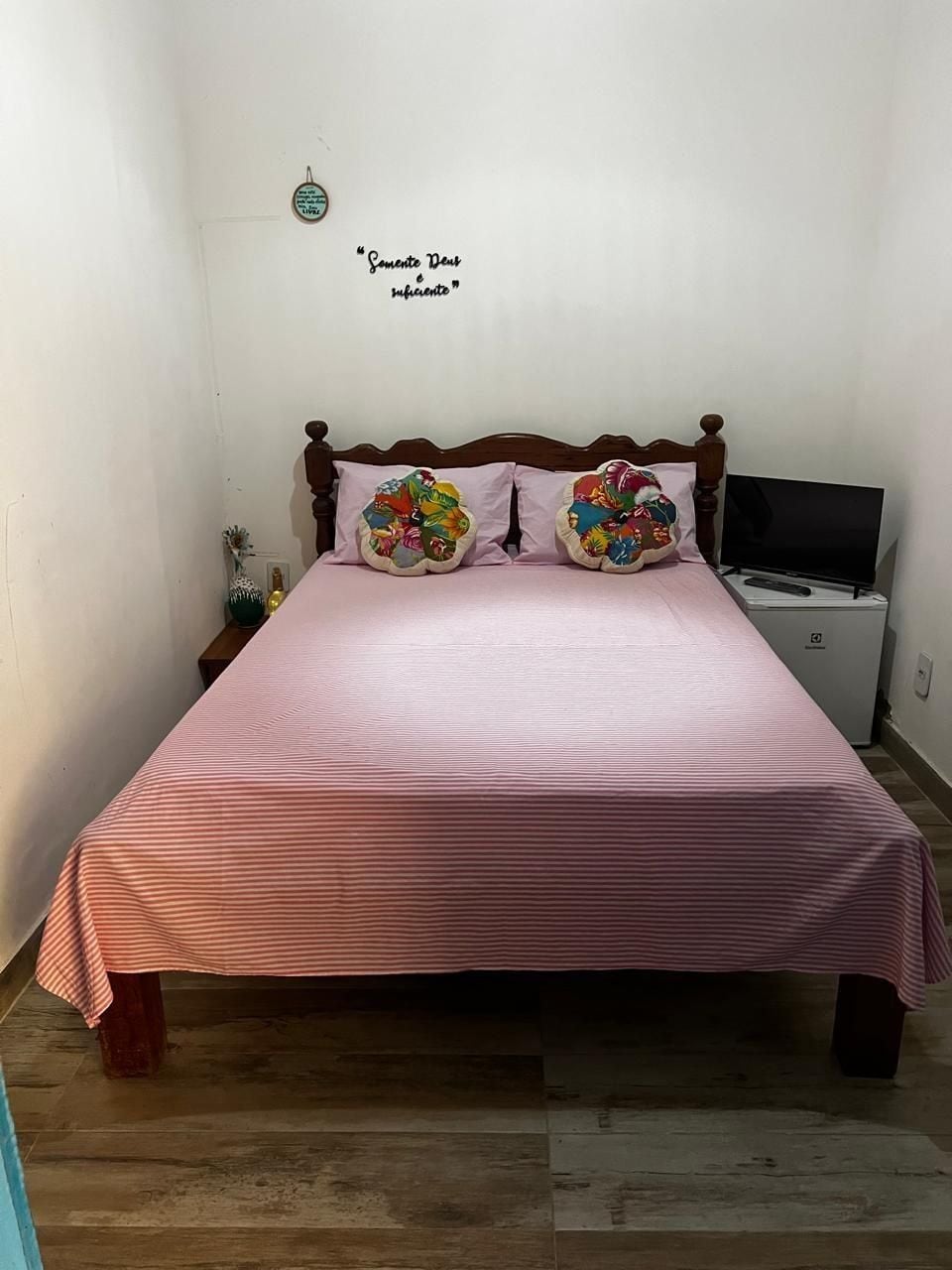 Uma cama com cabeceira de madeira, duas almofadas decorativas, um cobreleiro listrado em rosa, uma mesinha de cabeceira com um vaso e uma televisão sobre um suporte.