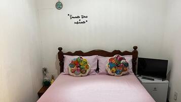 Uma cama com cabeceira de madeira, duas almofadas decorativas, um cobreleiro listrado em rosa, uma mesinha de cabeceira com um vaso e uma televisão sobre um suporte.