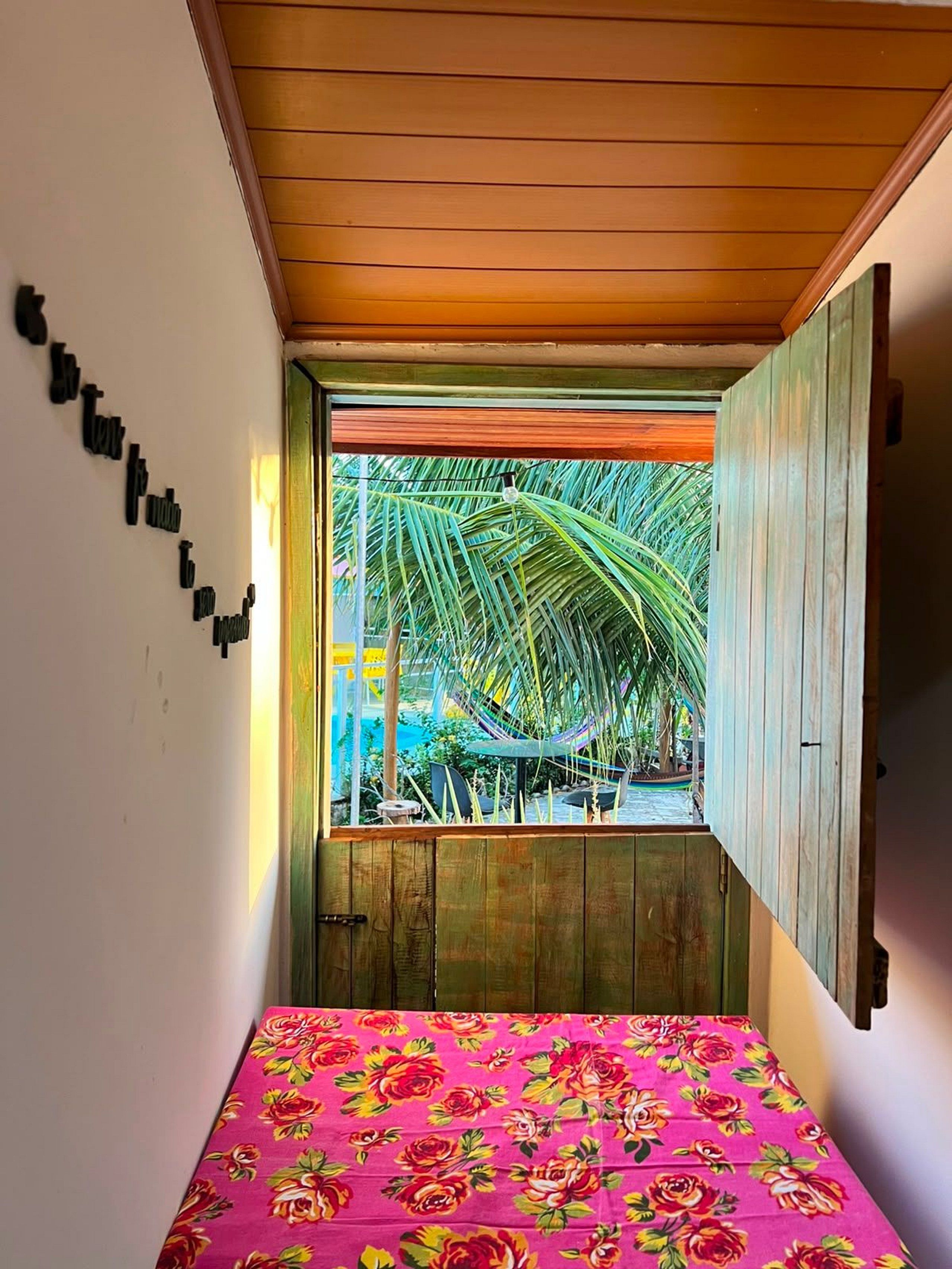 A room with a bed, a window with a view of palm trees, and a wooden ceiling.
