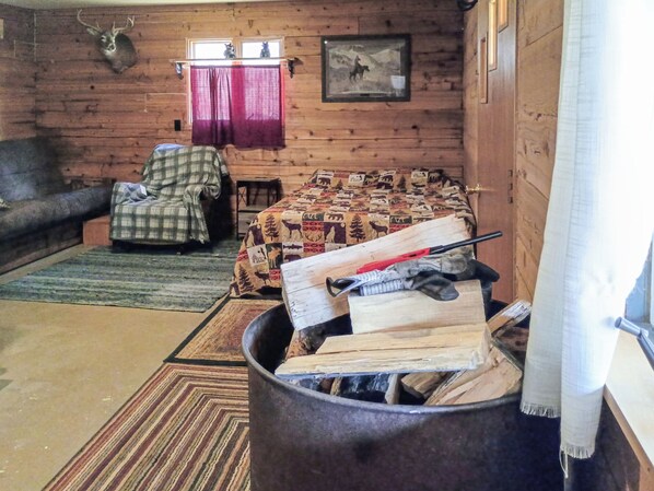 A cozy cabin interior with a plaid armchair, a bed with a forest-themed quilt, a fireplace with logs, and a mounted deer head.