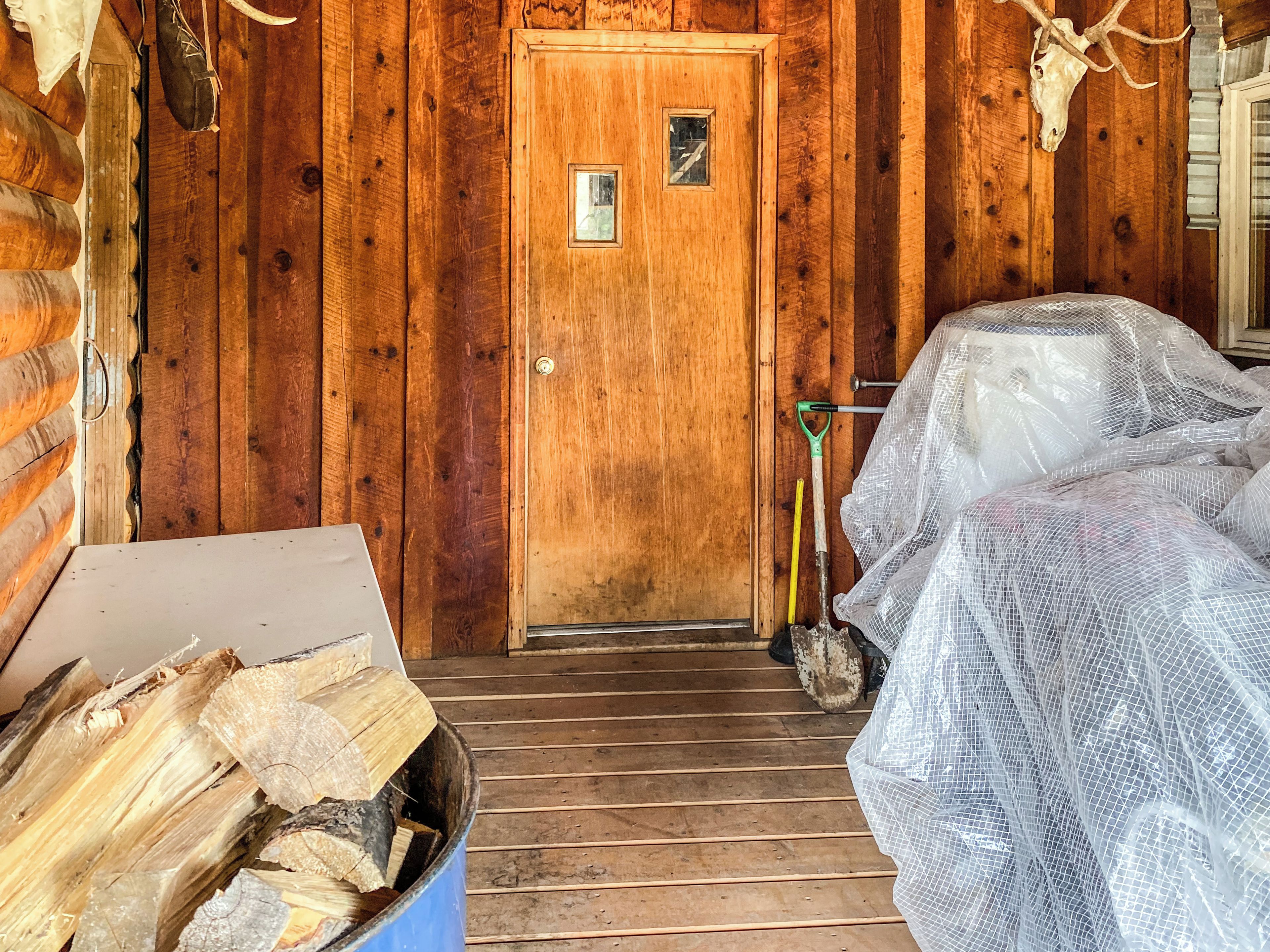 A wooden cabin interior with a door, a bed covered in plastic, and a box of firewood.