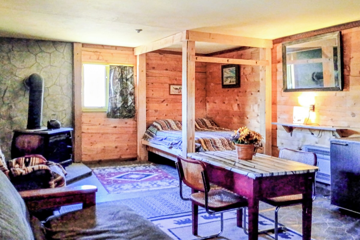 A cozy cabin interior with a bed, a table, chairs, a fireplace, and a stone wall.
