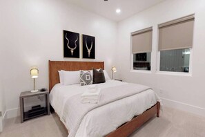 Ein Schlafzimmer mit einem Bett, zwei Nachttischlampen, einem Kopfteil, einem Fenster mit Jalousien und zwei an der Wand hängenden Bildern.