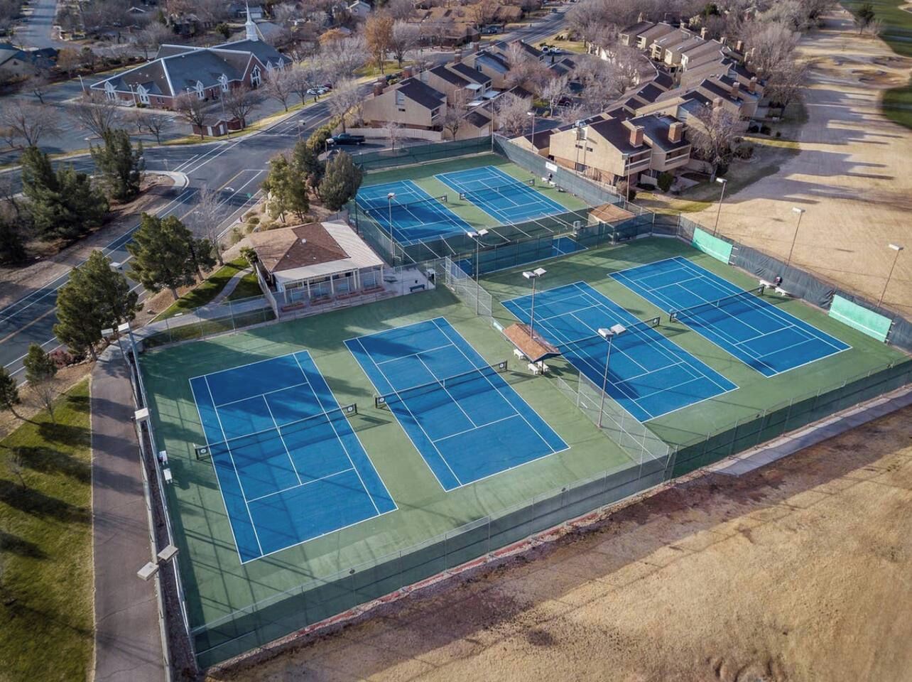 An aerial view of a tennis court complex with multiple courts, surrounded by residential buildings and a parking area.
