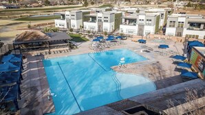 Une piscine entourée de chaises longues et de parasols, elle-même bordée de bâtiments modernes offrant une vue sur un paysage vallonné.