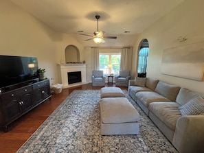 A living room with a television, fireplace, sofa, ottoman, and patterned rug. - Cedar Lake Cottage • Pool & Lake view! (Oxford)