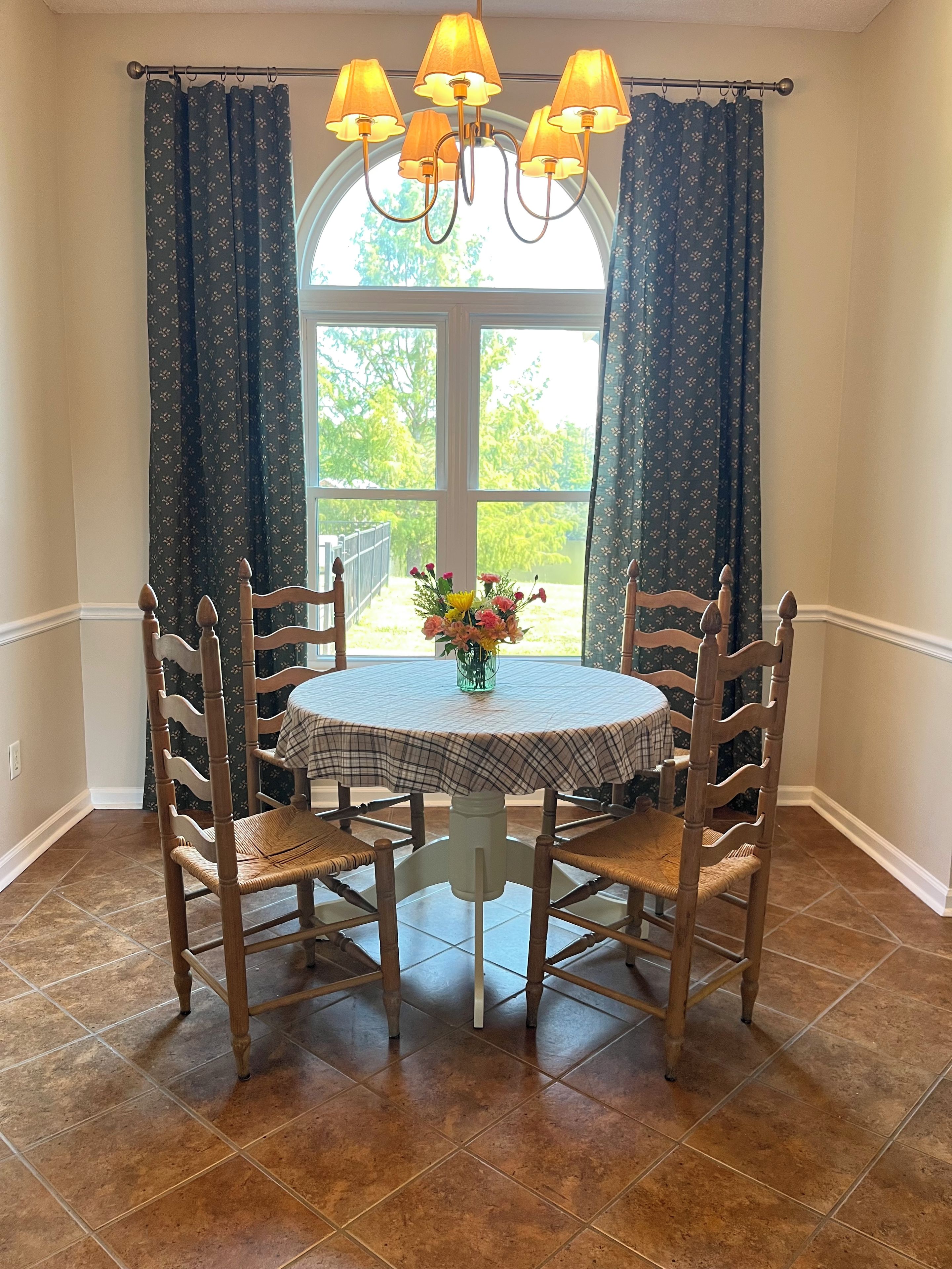 A dining area with a round table, four wooden chairs, a chandelier, and a window with curtains.