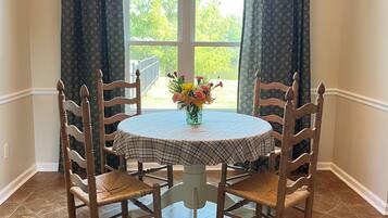 A dining area with a round table, four wooden chairs, a chandelier, and a window with curtains.