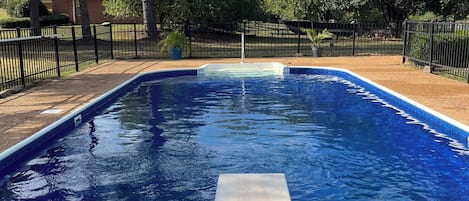 A rectangular swimming pool with a clear blue water surface, surrounded by a black metal fence, and a paved walkway leading to the pool.