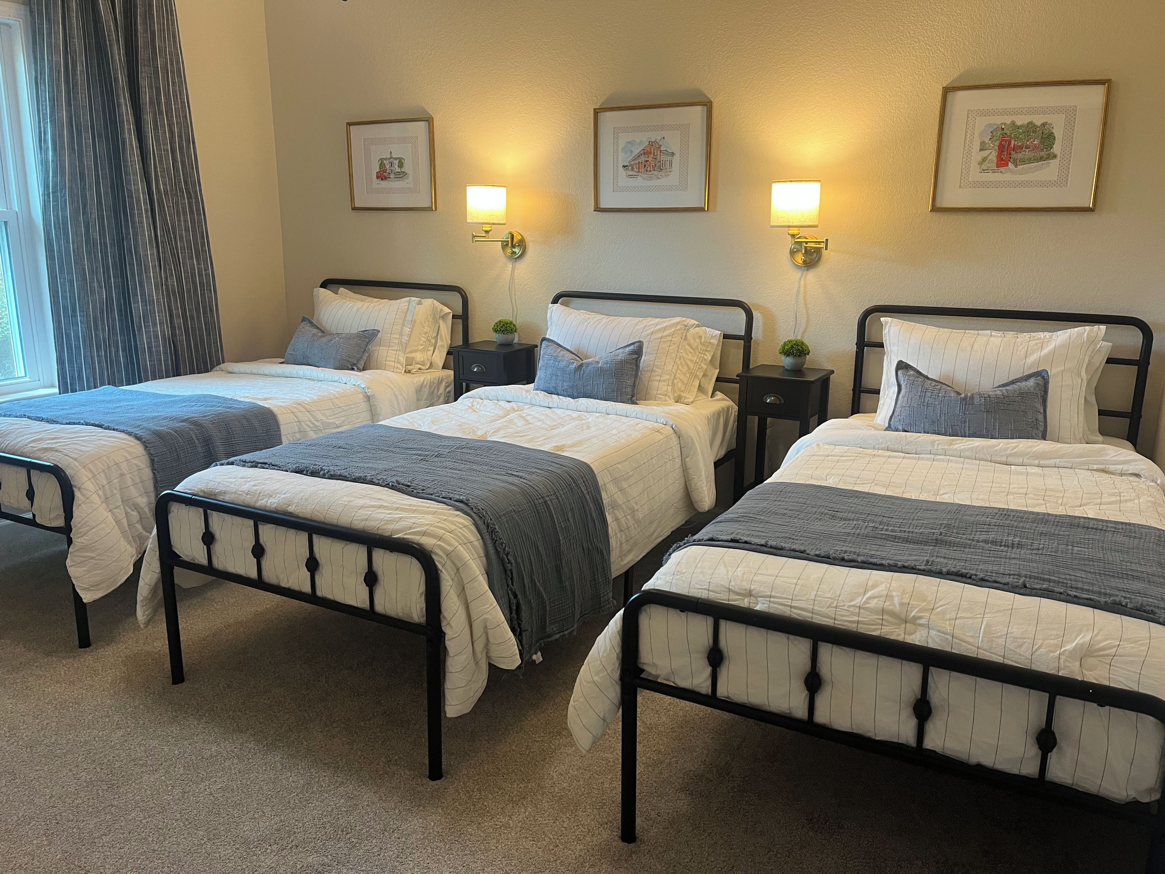 Une chambre d’hôtel avec trois lits, chacun doté d’un cadre en métal noir et de draps blancs. Des lampes murales et des tableaux encadrés ornent les murs.