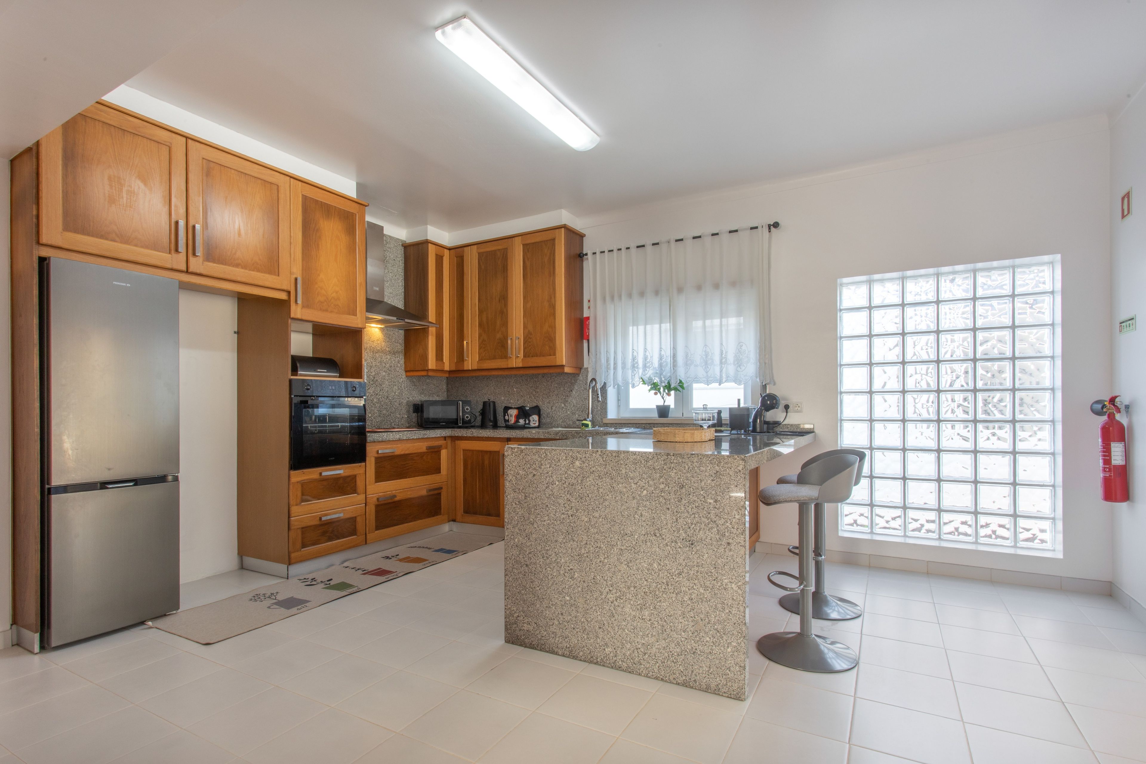 A modern kitchen with wooden cabinets, a stainless steel refrigerator, a built-in oven, and a granite countertop island with bar stools.