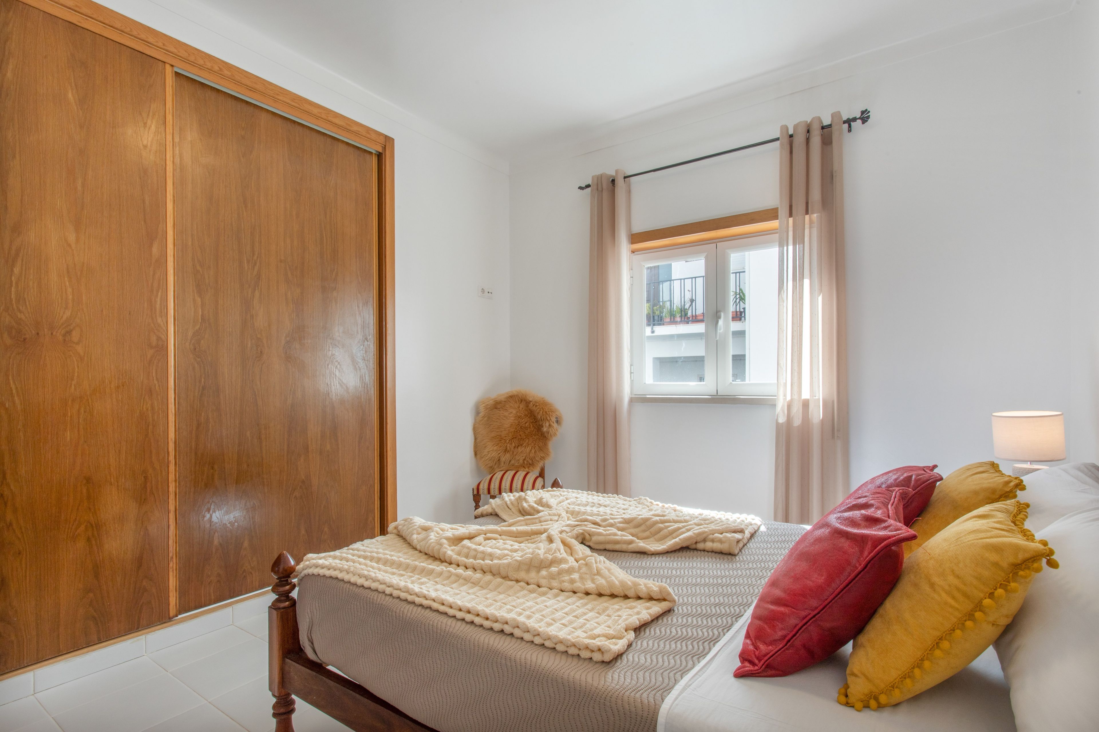 Un dormitorio con un armario de madera, una cama con colcha color beige, una ventana con cortinas y una lámpara.