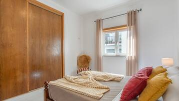 Un dormitorio con un armario de madera, una cama con colcha color beige, una ventana con cortinas y una lámpara.