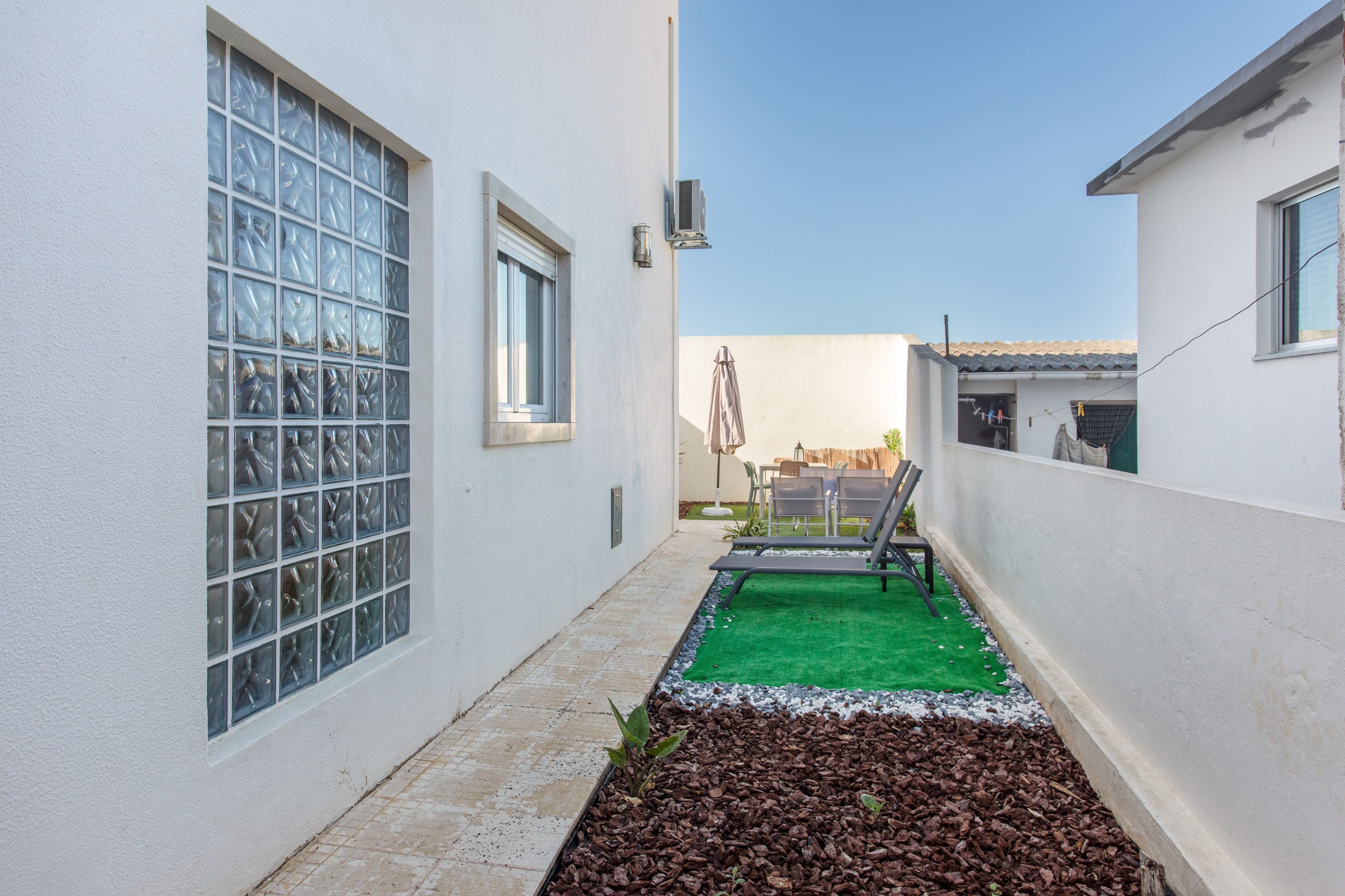 Un espacio exterior estrecho con área de césped artificial verde, una pequeña zona de estar con mesa y sillas, y una ventana con cuadricula de vidrios.