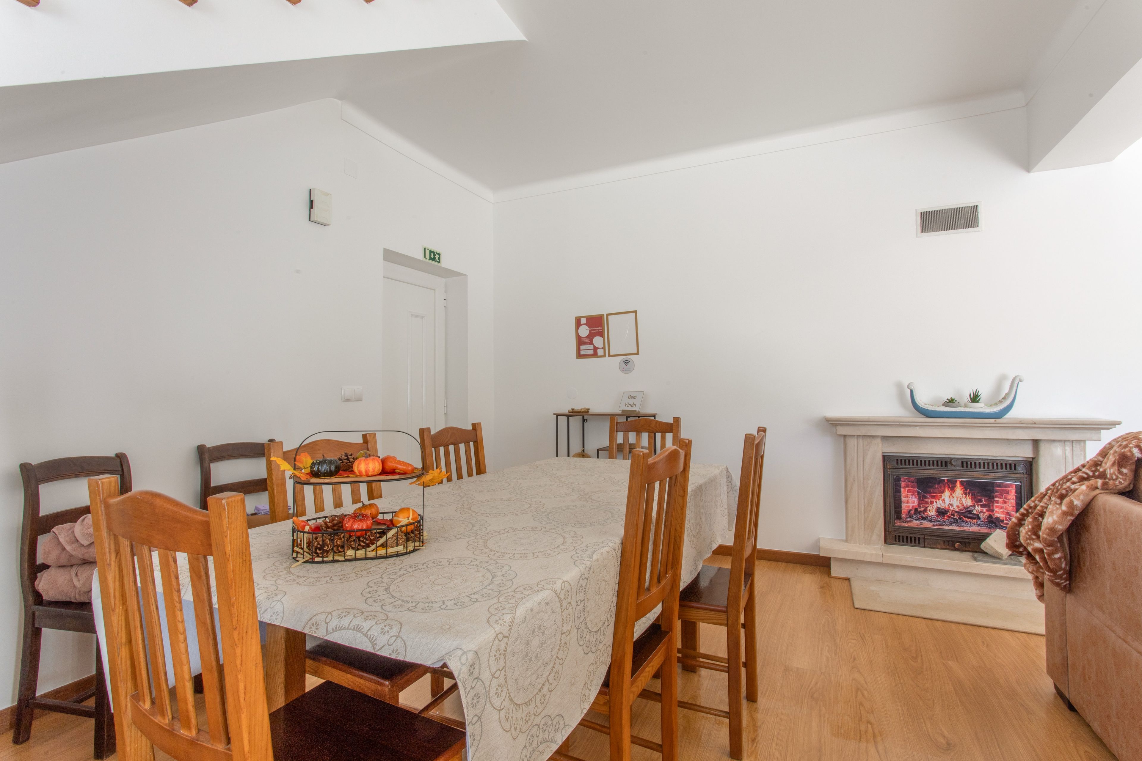 Uma sala de jantar com mesa posta para seis pessoas, rodeada por cadeiras de madeira. Um lareira com manto decorativo está visível à direita.