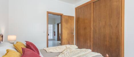 Een slaapkamer met een bed, kussens, een bedlampje en een houten garderobe.