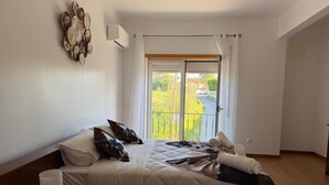 Un dormitorio con cama, almohadas, una lámpara de noche y vista a la calle a través de una ventana.