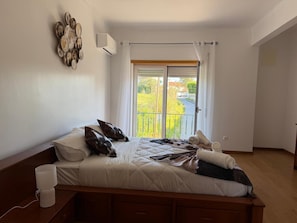 Un dormitorio con cama, almohadas, una lámpara de noche y vista a la calle a través de una ventana.