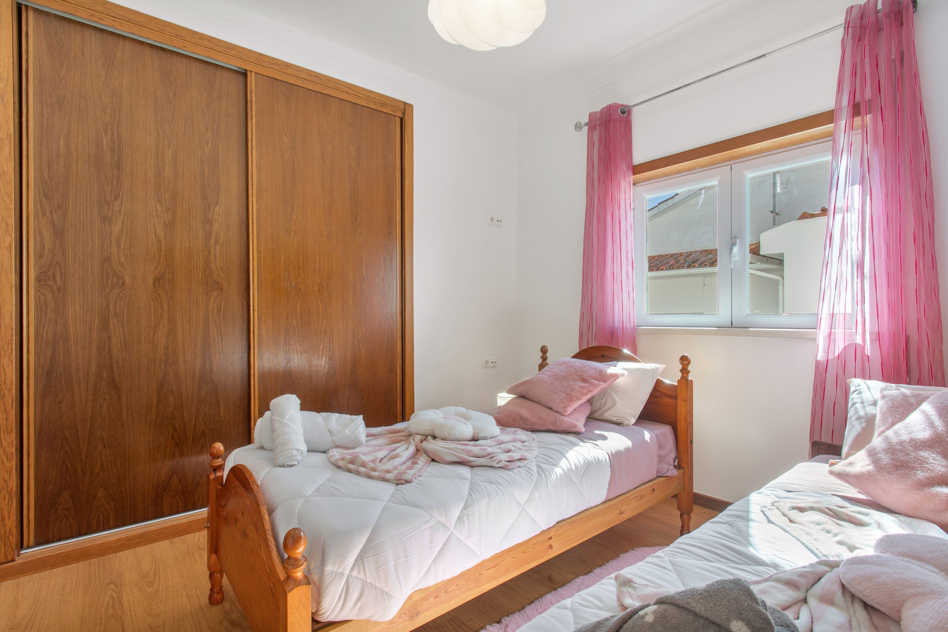 Quarto com guarda-roupa de madeira, cama com roupa de cama rosa e branca, e janela com cortinas rosa.