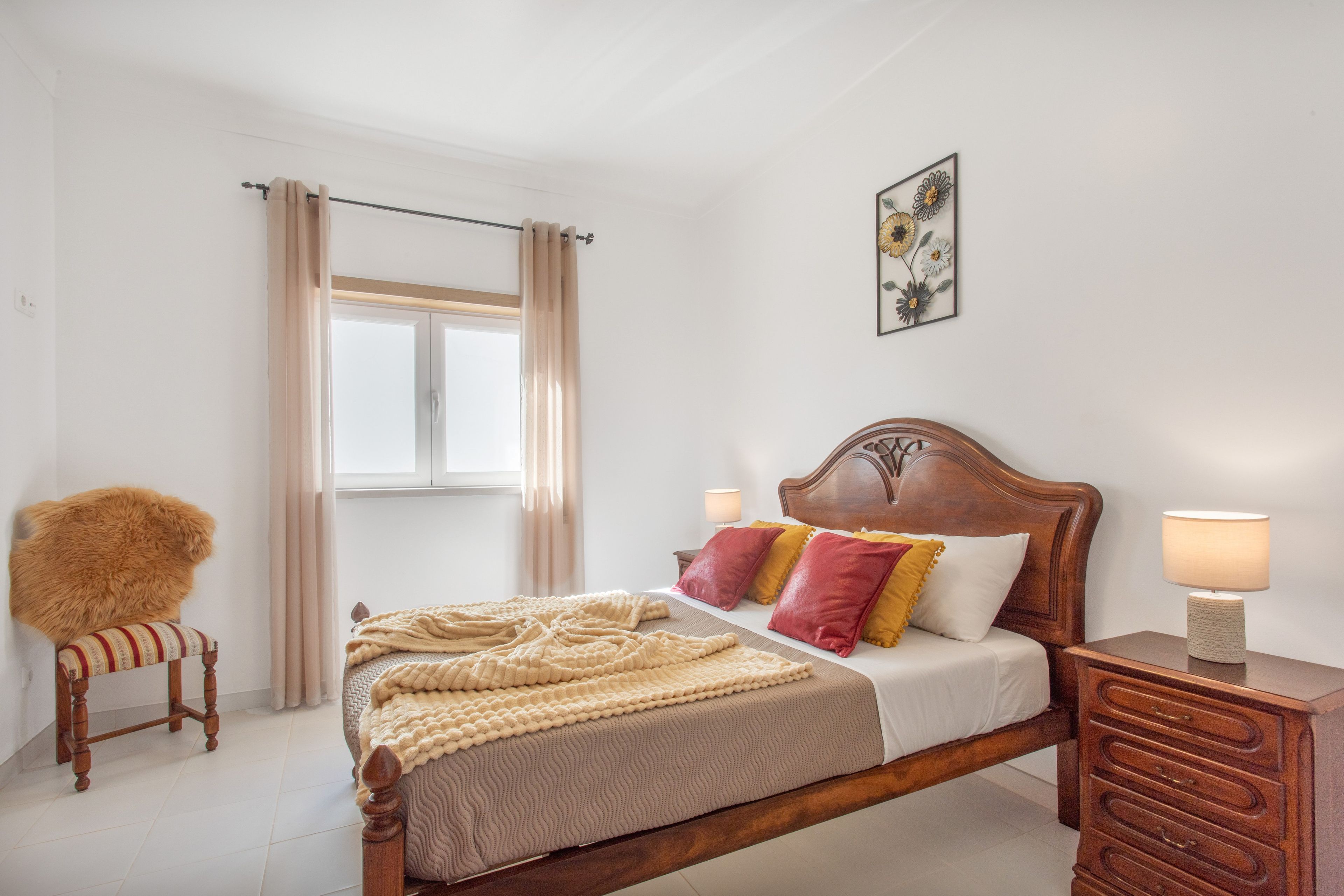A bedroom with a wooden bed, a nightstand, a chair, a window with curtains, and a wall-mounted picture.