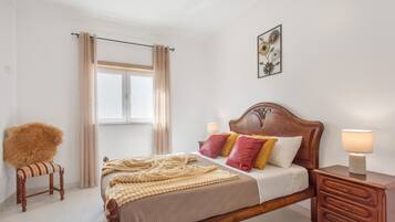 A bedroom with a wooden bed, a nightstand, a chair, a window with curtains, and a wall-mounted picture.