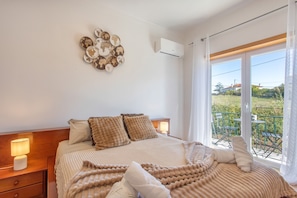 Un dormitorio con una cama grande, dos mesitas de noche, aire acondicionado instalado en la pared y vistas al exterior a través de una ventana.
