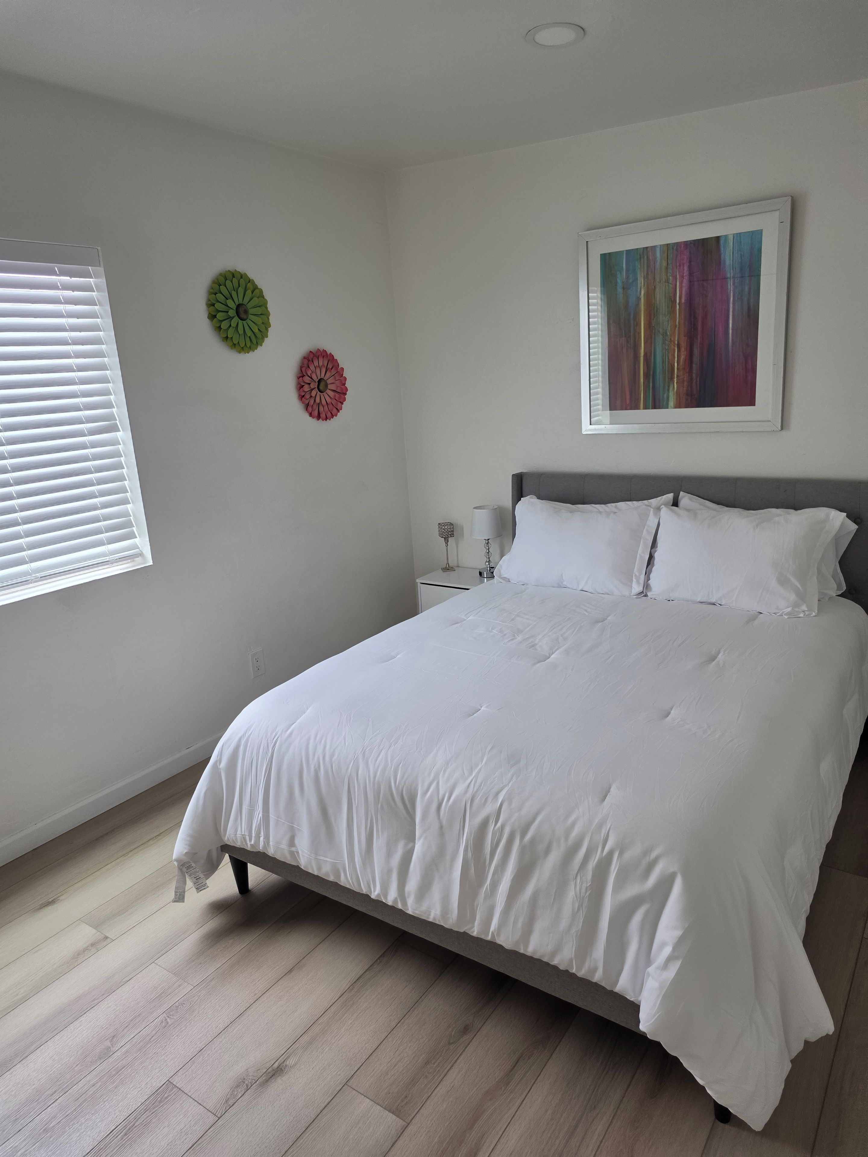 Une chambre à coucher comprenant un lit, une table de chevet, une lampe, une fenêtre avec des stores et deux éléments de décoration murale.