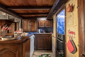 A rustic kitchen with wooden cabinets, a tiled backsplash, and a stone wall. - Hostcare - Le Petit Château – Downtown (Sarlat-la-Canéda)