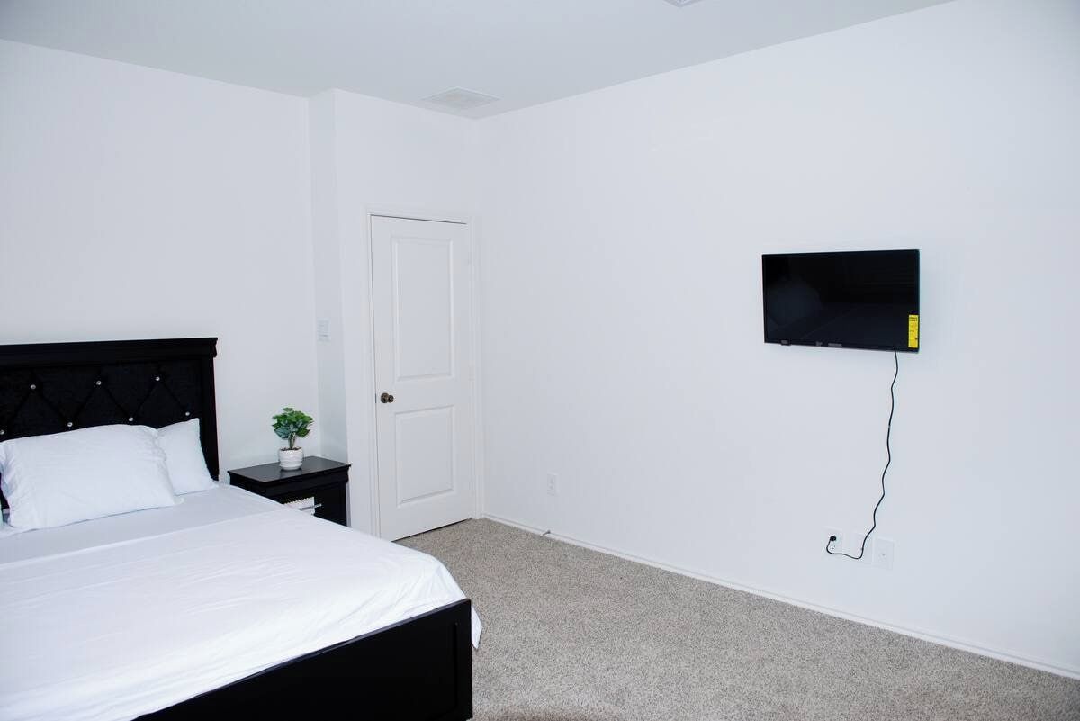 Un dormitorio con cama, una mesita de noche con una planta, una puerta blanca y un televisor montado en la pared.